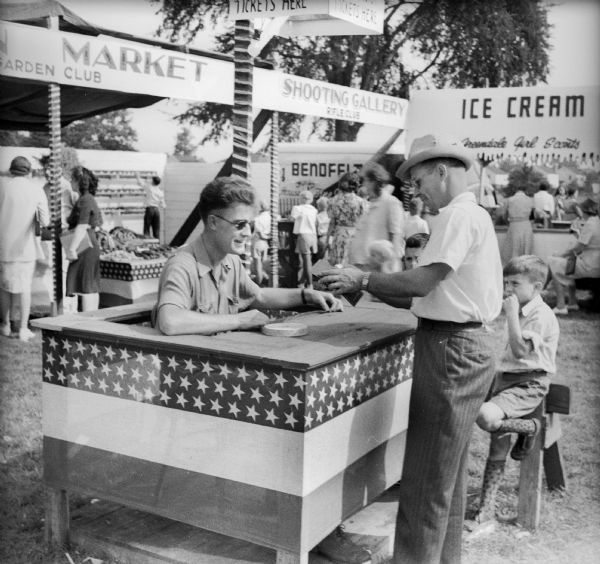 A man wearing eyeglasses and sitting at a booth sells tickets related to a Civilian Defense Rally. Children and adults are at booths in the background, including an ice cream stand.