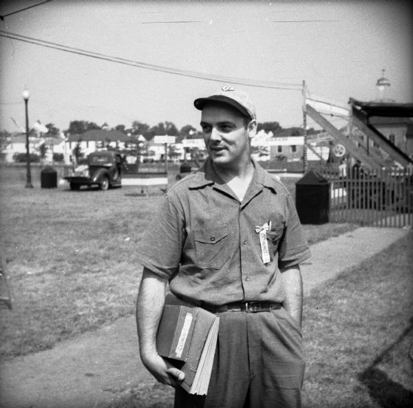 A man wearing a baseball cap holds a folder and a box labeled "Committee." He is also wearing a badge labeled "Committee" on his shirt pocket. In the far background are houses, and the Greendale Village Hall with clock tower.

James F. Ircink moved to Greendale in 1938 and resideed on Beaver Court. He was a village trustee from 1943-44 and from 1948-49. 