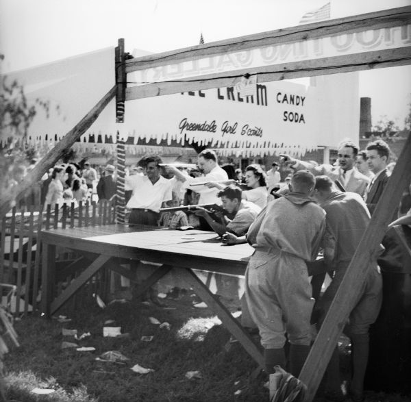 Side view from behind the shooting gallery at a Civilian Defense Rally. Women and boys are shooting at targets while Boy Scouts and other men stand around. The ice cream booth is behind them.