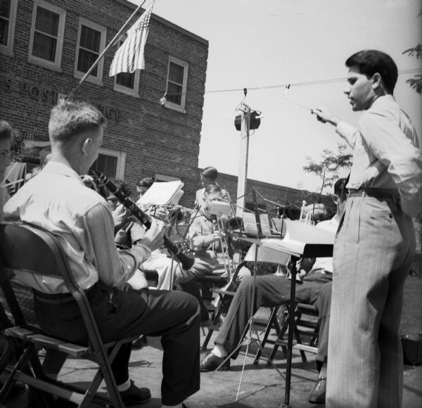 A conductor leads a band made up of school-age children as well as a few adults at a Civilian Defense Rally. They are playing outdoors in front of the Post Office.