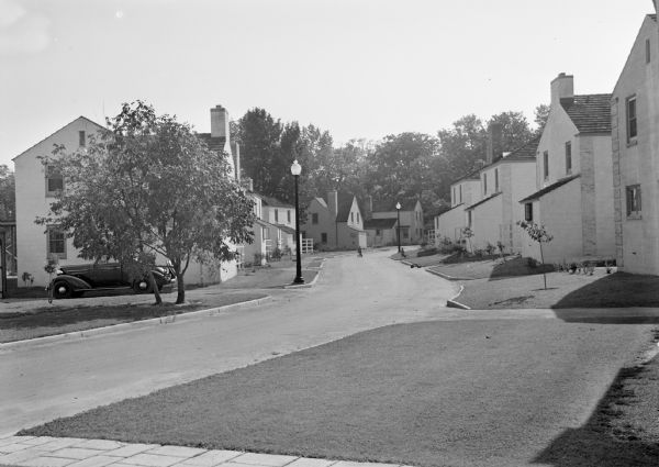 View from lawn of curved residential street lined with recently constructed housing and lampposts. A car sits in a driveway of a house on the left, and further down the road children play on tricycles.