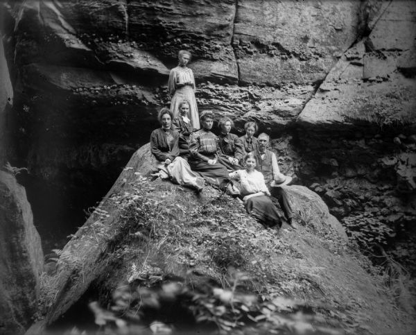 Seven women and a man pose on a large boulder in front of a wall-like rock formation, probably in Parfrey's Glen.