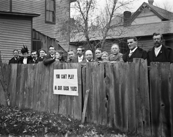 A group of adults, including Harry E. Cole (center), stand behind a fence. A sign on the fence reads, "YOU CAN'T PLAY IN OUR BACK YARD."