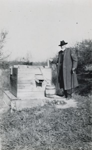 J.R. Hastie stands beside a chain and crank operated pump at the pioneer well, "a few miles below Portage."
