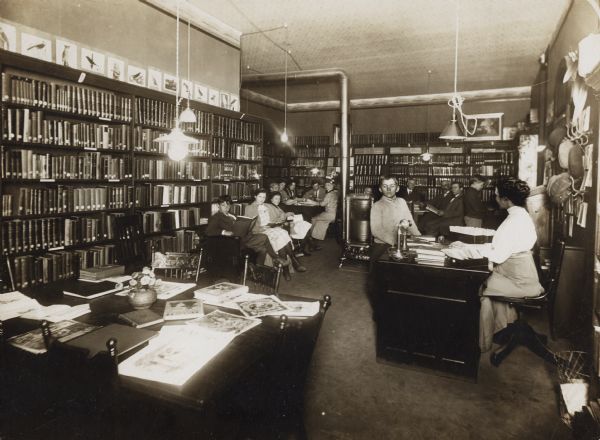 Interior view of the Brodhead Public Library. The back of the cardboard mounting reads: "Free Public Library, Brodhead, Wis." There is a stove in the center of the room. A woman (librarian?) is seated at a librarian's desk, and there are hat hooks and hats on the wall. Bookshelves line the walls, and a group of children and adults are using the room.