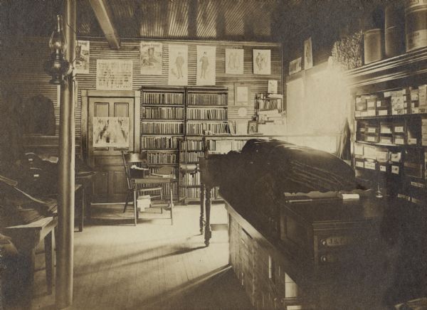 Interior view of the Genoa Junction Free Public Library. Reverse of cardboard mounting reads: "Genoa Junction Free Public Library, Genoa Junction, Wisconsin, Mrs. Carrie L. Manor Librarian. October 7, 1905." The library appears to be located in a dry goods store. There are bookshelves on the back wall, near a chair, small desk, and what appears to be a small card catalog box. On the right wall are shelves with labeled boxes, and in the foreground is a table with blankets.