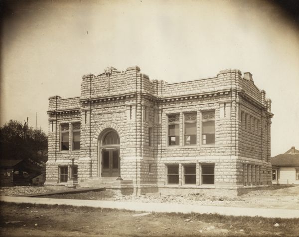 Exterior view of the Sturgeon Bay Public Library. Library opened in 1913 with a gift of $12,500 from Andrew Carnegie. Above the main entrance it reads: "Free Library." The stone building has an arched entrance. There is construction debris and unfinished landscaping around the building.

