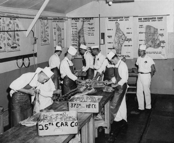 At Fort Sam Houston students from the Food Service School learn new meat cutting practices so that individual messes can pick up their rations ready for cooking. Prior to 1951 each mess had to cut up the carcasses. Posters along the walls describe beef cuts, and pork cuts.