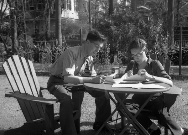 Harold Gauer and Robert Bloch sit on chairs outside in a backyard writing on sheets of paper. A liquor bottle sits on the table between them.