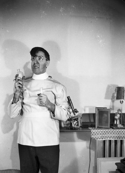 Harold Gauer in costume as a mad scientist. He is holding laboratory glassware, and a microscope is visible on the shelf behind him.