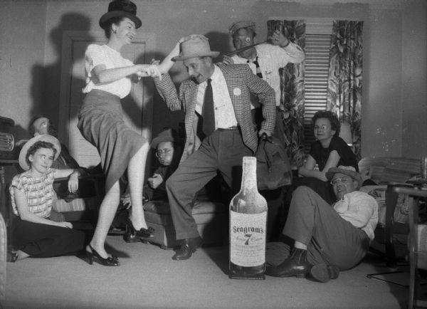 A couple dances behind an oversized cut-out of a bottle of Seagrams Whiskey, with other people seated around them. Some are wearing fake eyes.