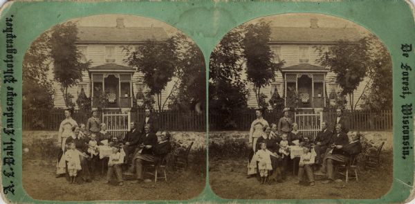 A family of twelve is grouped outdoors around a table. Behind them is a picket fence and a frame house, with latticework and carpenter's lace decorating its small porch.