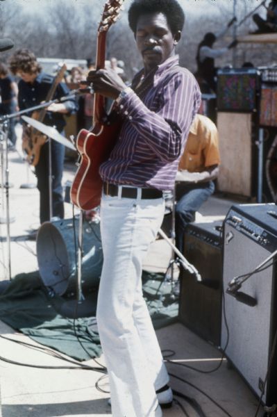 Guitarist Luther Allison on stage at the Sound Storm festival playing a Gibson guitar, with a drummer and bassist visible behind him.