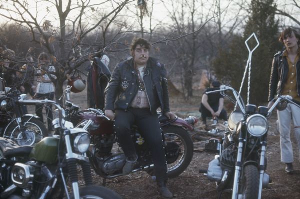 Two bikers with parked motorcycles. The biker on the left is wearing a large ring through his nose, a headband in his hair, a large swastika necklace, and leather jacket. He is seated on a Triumph motorcycle. The biker standing on the right is wearing a leather jacket and white pants.