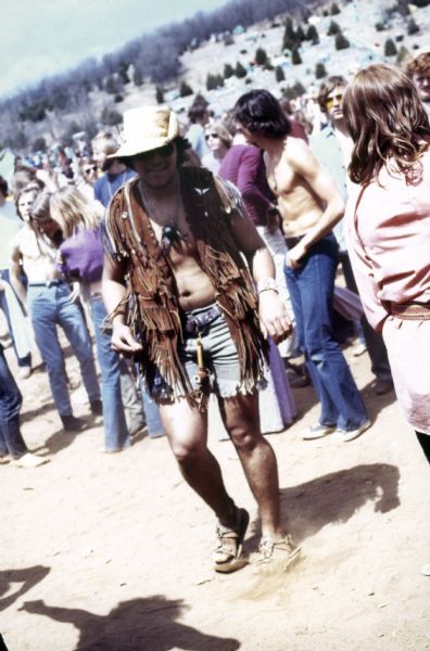A man wearing a hat, shorts, and fringed vest, with a pipe strung onto hit belt loop, dances among a crowd of other audience members at the Sound Storm music festival.