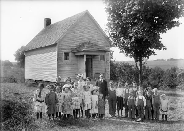 A group of children with their teacher posing on the lawn outside a one-room school building.