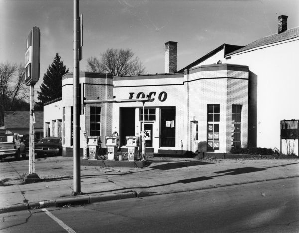 View from street of an old brick Ioco gas station with a few cars parked on the left.