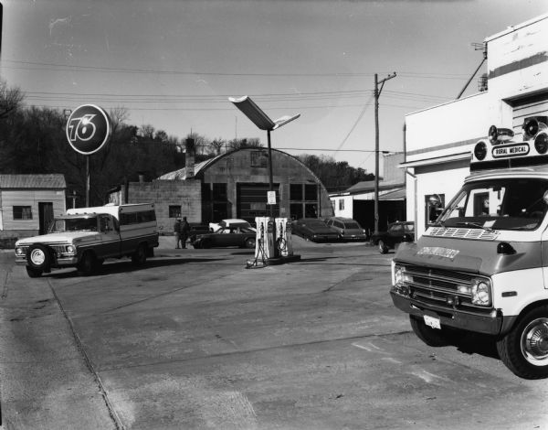 View across 76 Service Station parking lot towards a Quonset-hut style corrugated metal building across a street. An ambulance is in the right foreground, and other trucks and automobiles are parked on the street and in driveways.