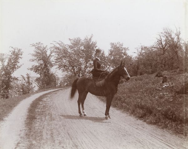 Mrs. Elsie Stevens poses on a path by a lake while riding a horse. Possibly on the University of Wisconsin-Madison drive (present-day Lakeshore path).