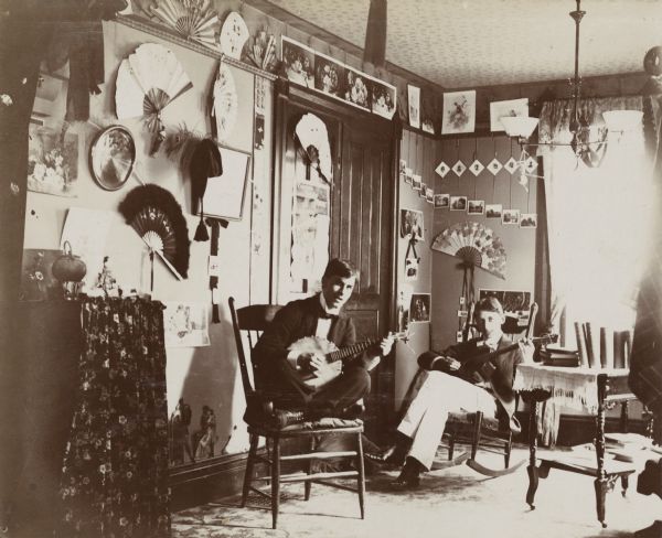 Two University of Wisconsin-Madison students, Mr. Rider and Mr. Broughton, sit in their residence at 625 Langdon Street and play musical instruments. Mr. Rider sits cross-legged on a chair with a banjo and Mr. Broughton reclines in a rocking chair with a guitar. There are a number of hand fans displayed on the walls.