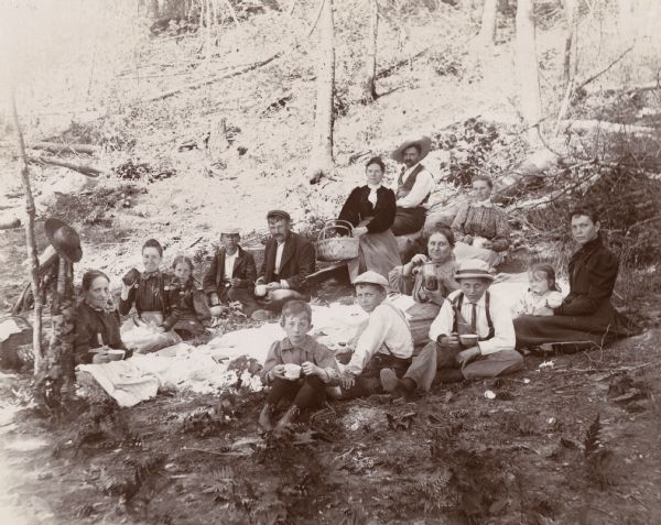 A group of men, women, and children gather for a picnic in the woods. A large picnic blanket covers the ground as the individuals pose for a photograph.