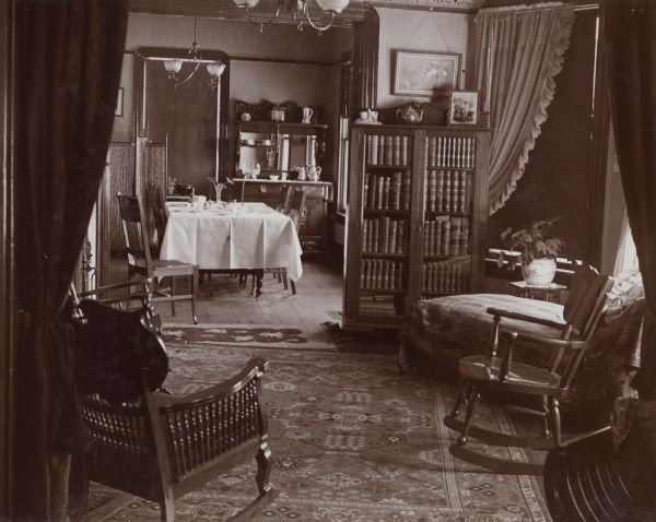 An interior view of the Wisconsin State Treasurer, Sewell A. Peterson, home on South Fairchild Street. The photograph is taken looking through the parlor and into the dining area. The Victorian-style interior includes patterned area rugs,  heavy draperies, and various pieces of wooden furniture, including a hutch with a beveled glass back.