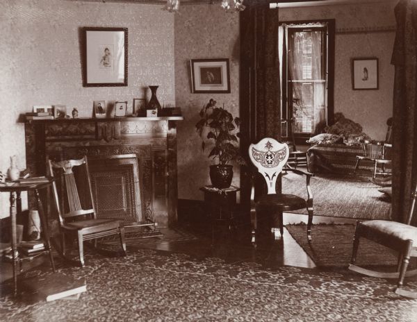 An interior view of Mrs. Hutchin's house. The Victorian-style interior consists of patterned wallpaper and area rugs, a fireplace, framed pictures and various styles of wooden furniture.