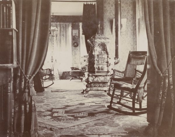 Interior view of the H. Schildhauer home at 13 North Webster Street. The photograph looks into a living area with a large, wood-burning furnace and several rocking chairs. Beyond the living room, another sitting room is visible through a large curtained doorway.