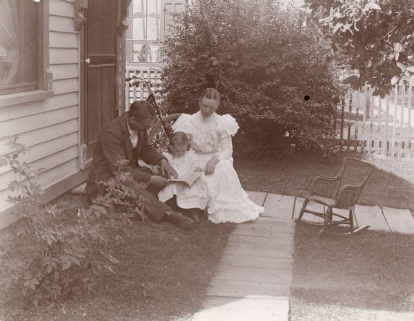 F.D. Eyerley sits with his wife and his daughter on the front stoop of their home on West Johnson Street. They are reading from a book. A small, wicker rocking chair is near the front walk.