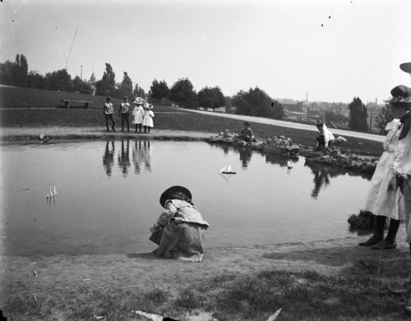 Syl playing with toy boats at a small pond at Reservoir Park. Other children and adults are around the pond which is on a hill. In the background below the hill are the spires of churches and roofs of houses.