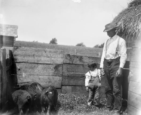 Syl with Uncle Herman who is wearing a hat. They are standing in a pig pen with four pigs eating in a corner.