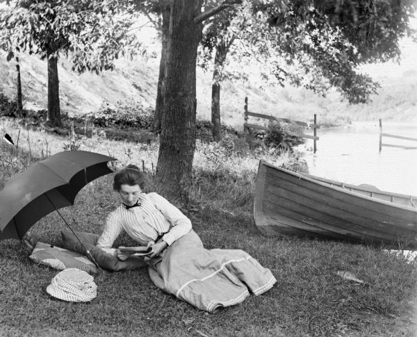 Woman lying in the grass reading near an umbrella along a shoreline. A boat is beached on the right, and in the background is a fence and hill.