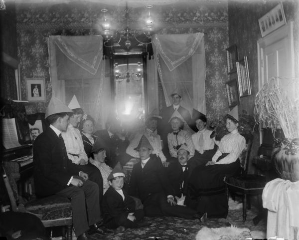 Men, women and children seated in chairs and on the floor wearing paper hats in honor of aunt Helen's birthday. A piano is on the right, a chandelier hangs from the ceiling, and a mirror and framed pictures are on the walls.