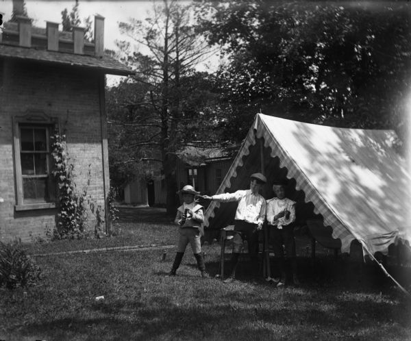 Three boys holding fireworks stand near a tent or open-sided awning in a backyard near a house.