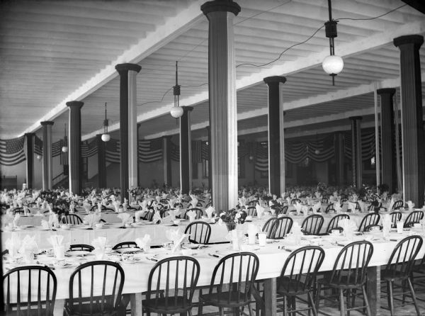 American flags drape the walls of a large empty hall prepared with one thousand place settings for soldiers returning from the Spanish-American War.