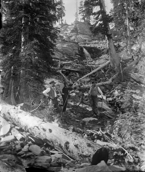 Three man stand in a small clearing on a steep rocky hill amid fallen trees. The small waterfall behind the men was the water power for the Leighton-Wyoming mining camp. One of the men stands behind a camera on a tripod. Joe Leighton is the mustachioed man on the far left.