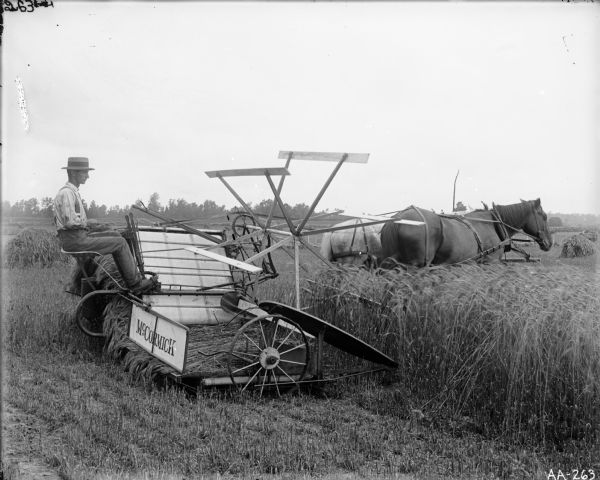 Young Man on Horse-Drawn McCormick Binder Side view of a young man wearing a braided and banded hat operating a McCormick horse-drawn grain binder in a field. Piles of grain are in the background.