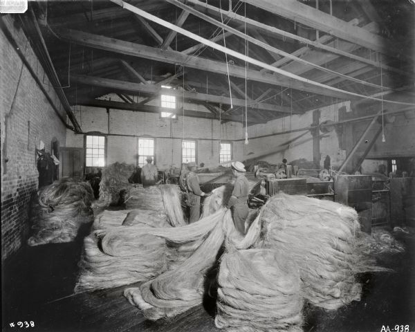 Men feeding fiber into manufacturing machines standing on a large factory floor with a high ceiling, many exposed ceiling beams, windows, and brick walls. Piles of coiled fiber are on the floor in front of machinery. The factory is possibly the McCormick Twine Mill.