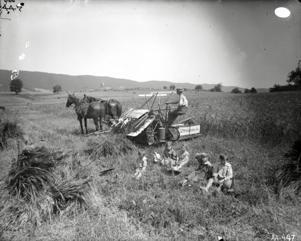 Elevated view of five children sitting in a field of grain. Behind them a man operates a McCormick grain binder pulled by two horses, one wearing a fly-net. In the background is a fence, and beyond is a range of hills.