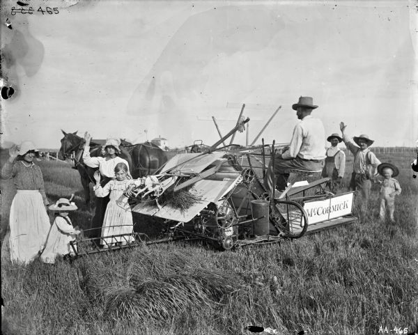 A man with his back turned is posing on a horse-drawn McCormick grain binder in a field. On his left are four females, including a woman, one younger woman, and two girls, wearing hats and bonnets and waving at the photographer. On the right three males are standing — an adult, a youth, and a child, are also waving. Farm buildings and a fence are in the background.