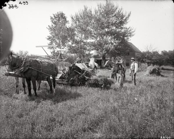 A man smoking a pipe sitting on a horse-drawn grain binder. Around him are young boys with horses. Another man is standing on the right. In the background is a farmhouse.