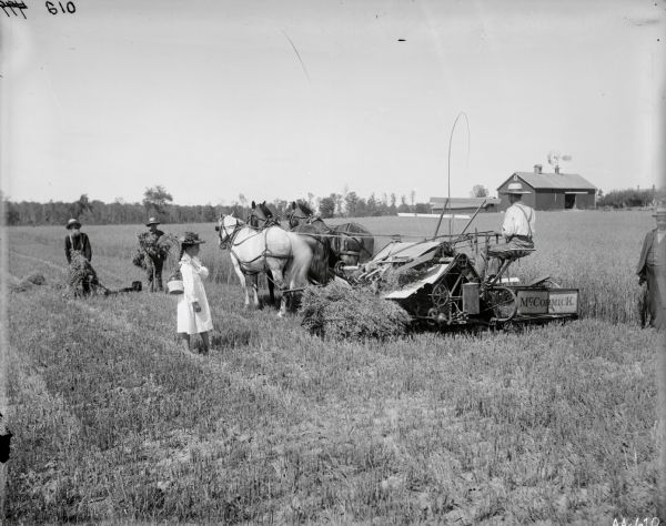 A group of people are posing in a field near a barn. They are harvesting grain, and a man is operating a grain binder pulled by three horses. A girl wearing a hat and dress is standing in the field with a jug over her shoulder. Two men are holding armfuls of grain. Another man wearing a suit and hat is standing on the right.