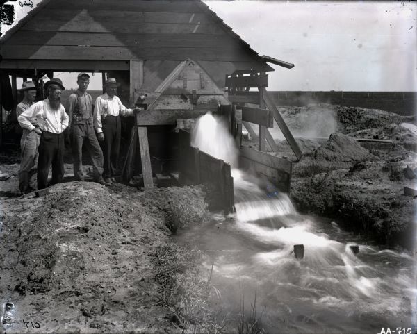 Men are standing next to a wooden structure, probably a pump house for an irrigation system. Water is streaming from the structure into a channel while the men are looking on.