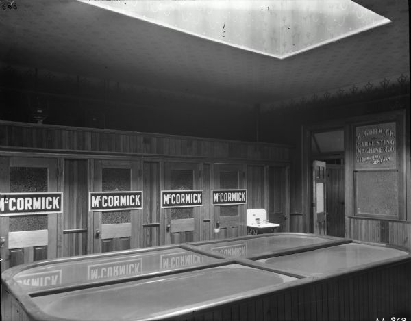Interior of a McCormick agency or dealership building owned by A.I. Dourgherty (as written on the glass of the window). A group of four bathtubs are in the center of the room under a skylight, and behind them are several doors with "McCormick" signs over them. A sink is on the right near the door.