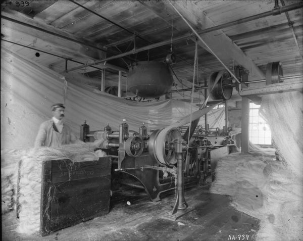 A factory worker uses a spreader machine to work with sisal fiber during the production of binder twine at the McCormick Twine Mill. White sheets are hanging in the background as backdrops.