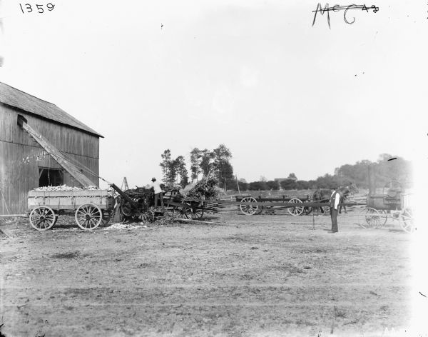A man working with wagons, an engine, and a husker-shredder near a barn, while another man is looking on. One wagon is filled with corn cobs.
