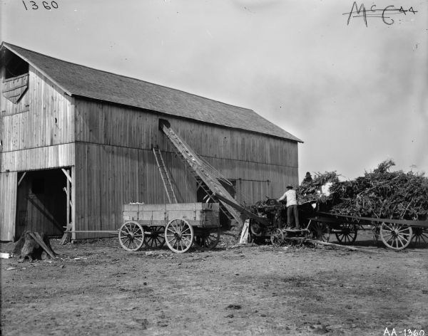 Men working with a husker-shredder and wagons near a barn.