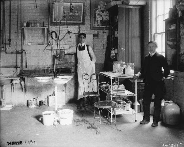 A doctor(?) and a young man standing in a factory medical office or first aid station, most likely at McCormick Works. Pails and jugs are on the floor, and bottles and canisters are arranged around the room. Two advertising posters are hanging on the wall, including on called "Back from the War."