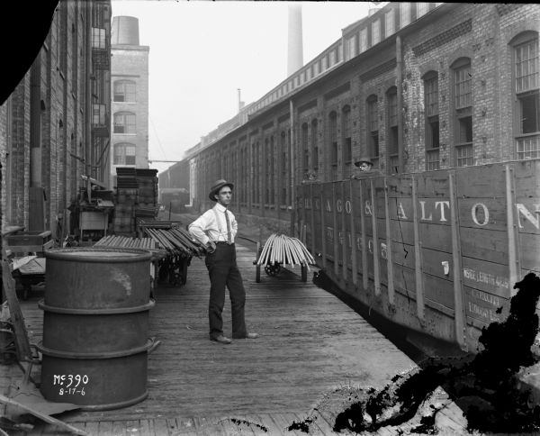 A man is standing on a railroad loading dock near a factory building, probably the McCormick Works. He is dressed in dark pants, a light-colored shirt, suspenders, and a hat. On the dock is a large barrel and what appears to be several carts of building or manufacturing supplies. On the railroad tracks are a boxcar for the "Chicago & Alton" railroad company. Two men inside the boxcar are leaning against the inside of the car, with only their arms and heads visible above the sides.