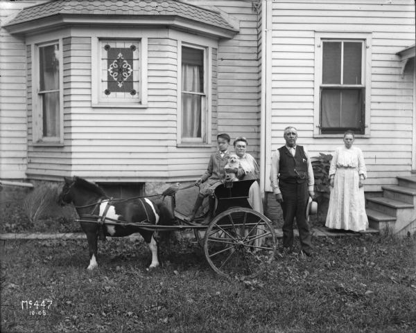 A boy dressed in a light suit and dark socks is sitting in a buggy pulled by a miniature horse. In the boy's arm is a small dog, and a parrot is perched on the reins. Behind the boy are three adults, two women are dressed in light-colored skirts and blouses, and a man is dressed in dark pants, a dark vest with a pocket watch, a light-colored button-up shirt holding a light-colored hat. The house in the background features a stained glass window. Vicinity of Chicago.
